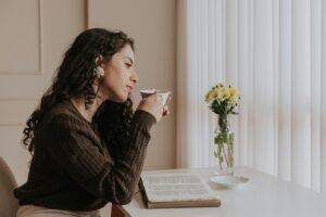 A woman sitting by a window at home, calmly drinking tea beside an open book and a vase of flowers, showing a peaceful daily home routine.