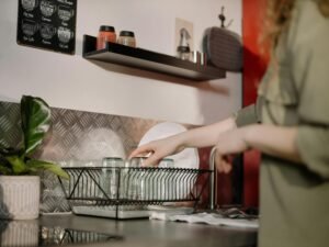 Person placing clean drinking glasses into a black dish rack on a kitchen counter, with shelves, jars, and kitchen decor visible in the background.