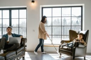 A bright, cozy living room where a woman cleans the floor while a man works on a laptop and a child relaxes on an armchair, showing everyday family life at home.
